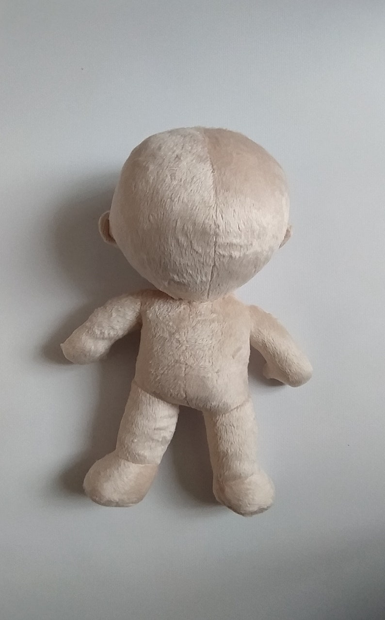Op de afbeelding: Een beige pluche pop met een groot hoofd en kleine ledematen. De pop staat op een wit oppervlak.