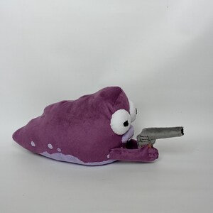 Puede incluir: Un peluche con forma de mancha morada con ojos blancos y negros, sosteniendo una pistola de juguete gris. El juguete tiene manchas blancas y una parte inferior morada m&aacute;s clara. El fondo es blanco liso.