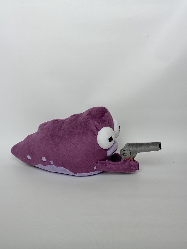 Puede incluir: Un peluche con forma de mancha morada con ojos blancos y negros, sosteniendo una pistola de juguete gris. El juguete tiene manchas blancas y una parte inferior morada m&aacute;s clara. El fondo es blanco liso.