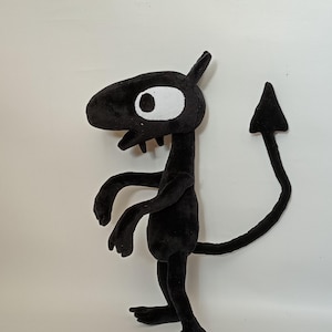 Puede incluir: Un juguete de peluche negro con forma de criatura, hocico largo, un solo ojo grande blanco con pupila negra y dos dientes blancos pequeños. La figura tiene una cola larga y delgada que termina en una punta triangular y se mantiene en pie.