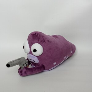Puede incluir: Juguete de peluche con forma de criatura alien&iacute;gena morada con grandes ojos blancos y una pistola de juguete gris. El peluche tiene manchas blancas y una boca de color lila claro.