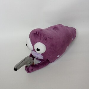 Puede incluir: Juguete de peluche con forma de alien&iacute;gena morado con ojos blancos grandes y una pistola de juguete gris. El alien&iacute;gena tiene boca y brazos de color morado claro. Lunares blancos en el cuerpo del alien&iacute;gena. El juguete est&aacute; sobre un fondo blanco.