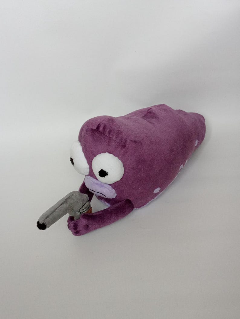 Puede incluir: Juguete de peluche con forma de alien&iacute;gena morado con ojos blancos grandes y una pistola de juguete gris. El alien&iacute;gena tiene boca y brazos de color morado claro. Lunares blancos en el cuerpo del alien&iacute;gena. El juguete est&aacute; sobre un fondo blanco.