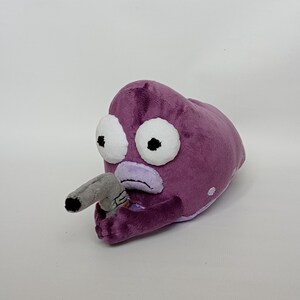 Puede incluir: Un peluche morado con forma de criatura con grandes ojos blancos y una nariz gris. La criatura sostiene un objeto gris en su mano. El juguete tiene una textura suave y difusa y est&aacute; sobre un fondo blanco.