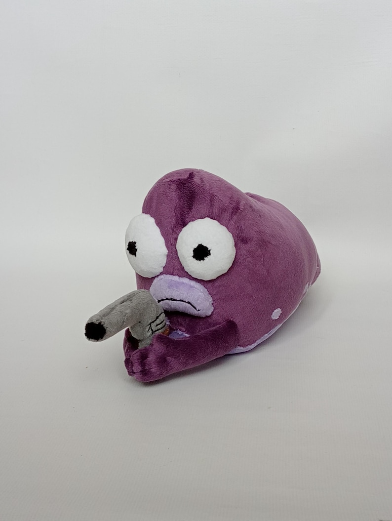 Puede incluir: Un peluche morado con forma de criatura con grandes ojos blancos y una nariz gris. La criatura sostiene un objeto gris en su mano. El juguete tiene una textura suave y difusa y est&aacute; sobre un fondo blanco.