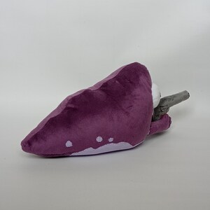 Puede incluir: Juguete de peluche morado con forma de criatura, con detalles blancos y un arma gris. El juguete est&aacute; hecho de material suave. El juguete est&aacute; sobre una superficie blanca.