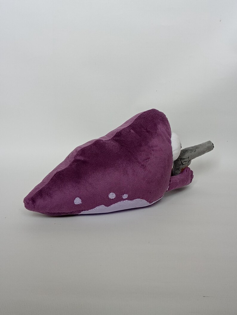 Puede incluir: Juguete de peluche morado con forma de criatura, con detalles blancos y un arma gris. El juguete est&aacute; hecho de material suave. El juguete est&aacute; sobre una superficie blanca.