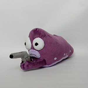 Puede incluir: Peluche morado con forma de criatura con grandes ojos blancos y una pistola gris. El peluche tiene una expresi&oacute;n triste y manchas blancas. El juguete est&aacute; hecho de tela suave.