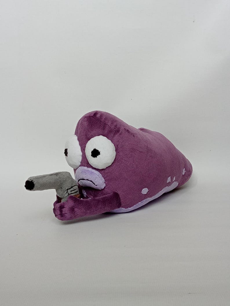 Puede incluir: Peluche morado con forma de criatura con grandes ojos blancos y una pistola gris. El peluche tiene una expresi&oacute;n triste y manchas blancas. El juguete est&aacute; hecho de tela suave.
