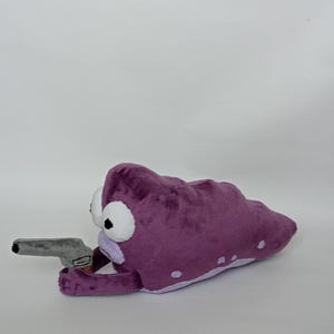 Puede incluir: Criatura de peluche morada con grandes ojos blancos y una pistola de juguete gris. El peluche tiene manchas blancas y una parte inferior de color p&uacute;rpura claro. El juguete est&aacute; sobre un fondo blanco.