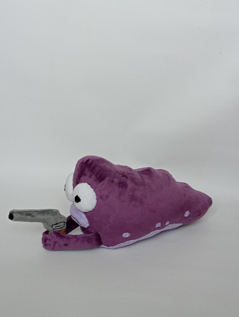 Puede incluir: Criatura de peluche morada con grandes ojos blancos y una pistola de juguete gris. El peluche tiene manchas blancas y una parte inferior de color p&uacute;rpura claro. El juguete est&aacute; sobre un fondo blanco.