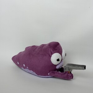 Puede incluir: Peluche con forma de criatura alien&iacute;gena morada que sostiene una pistola de juguete gris. La criatura tiene grandes ojos blancos con pupilas negras y una barriga de color lila claro. El juguete est&aacute; hecho de tela suave.