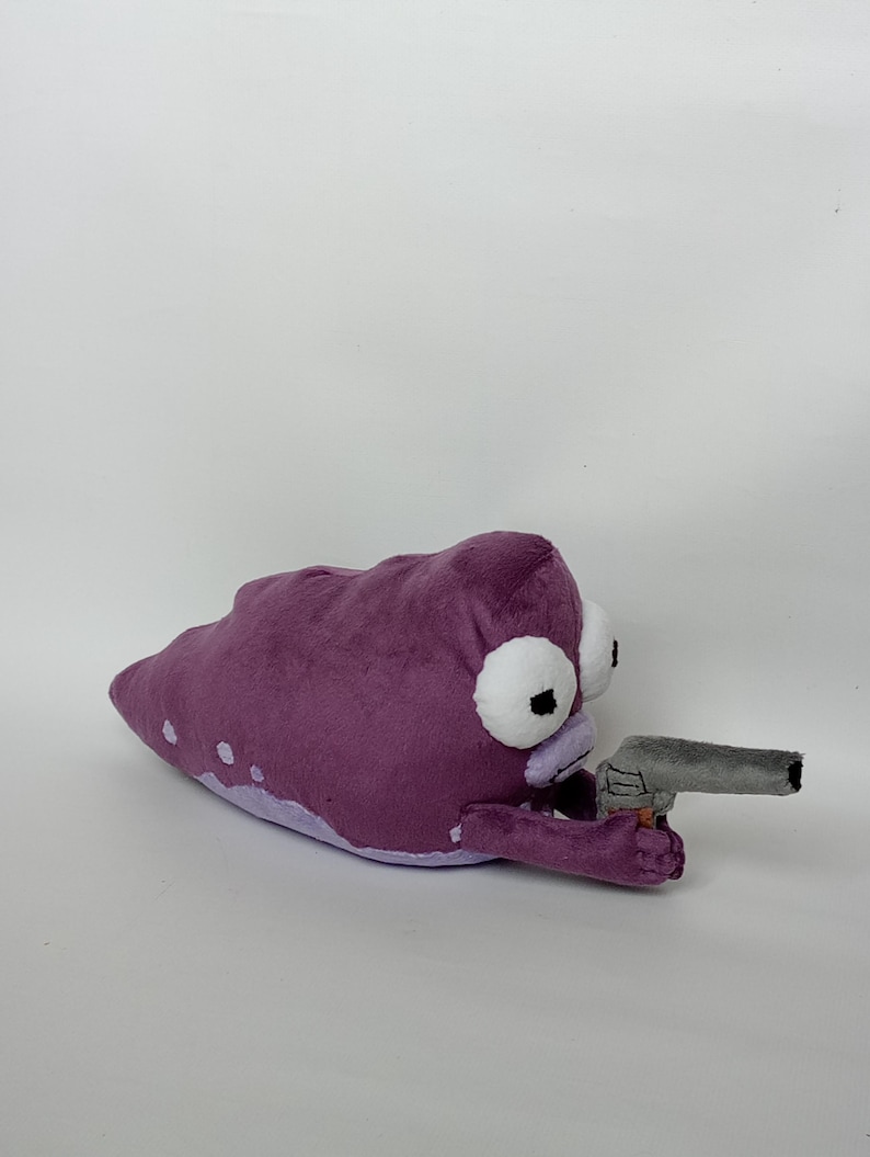 Puede incluir: Peluche con forma de criatura alien&iacute;gena morada que sostiene una pistola de juguete gris. La criatura tiene grandes ojos blancos con pupilas negras y una barriga de color lila claro. El juguete est&aacute; hecho de tela suave.