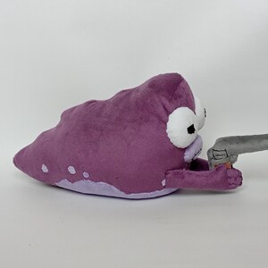 Puede incluir: Peluche morado con forma de mancha con ojos blancos y una pistola de juguete gris. El peluche tiene una parte inferior de color p&uacute;rpura claro y puntos blancos. El juguete est&aacute; sobre un fondo blanco.