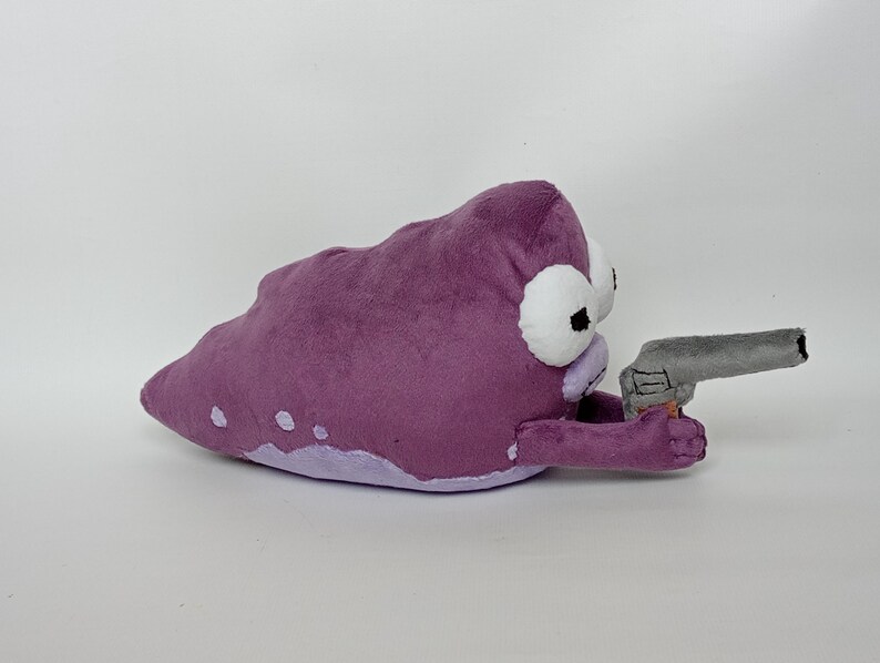 Puede incluir: Peluche morado con forma de mancha con ojos blancos y una pistola de juguete gris. El peluche tiene una parte inferior de color p&uacute;rpura claro y puntos blancos. El juguete est&aacute; sobre un fondo blanco.