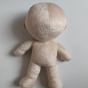 Op de afbeelding: Een beige pluche pop met een groot hoofd en kleine ledematen. De pop staat op een wit oppervlak.