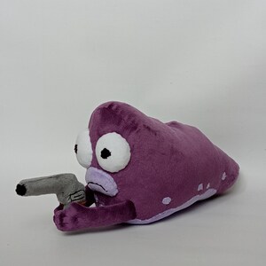 Puede incluir: Peluche morado con forma de criatura con grandes ojos blancos y una pistola de juguete gris. El peluche tiene un contorno morado claro y manchas blancas. La pistola de juguete es gris.