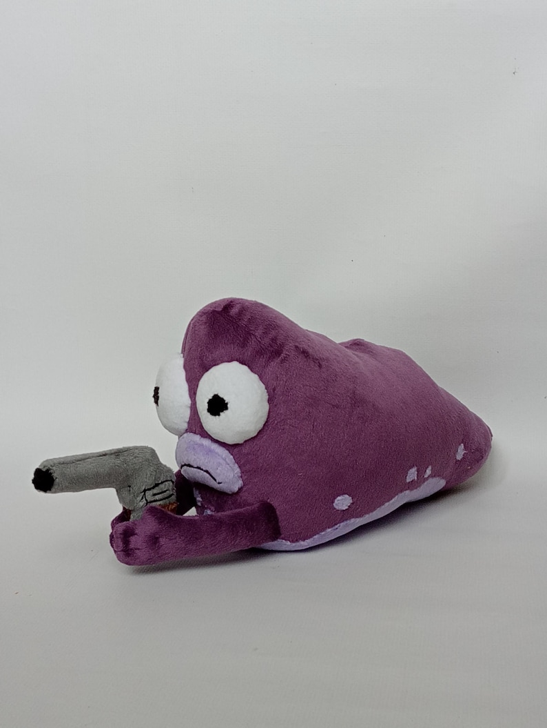 Puede incluir: Peluche morado con forma de criatura con grandes ojos blancos y una pistola de juguete gris. El peluche tiene un contorno morado claro y manchas blancas. La pistola de juguete es gris.