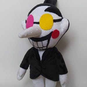 May include: A plush doll of a character with a white face, black hair, and a black suit. The character has a long white nose, a wide grin, and large eyes with pink, yellow, and red accents. The doll is standing upright.