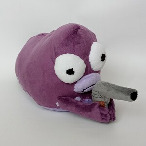 Puede incluir: Un peluche morado con forma de criatura con grandes ojos blancos y una pistola de juguete gris. El juguete tiene una textura suave y difusa, sobre un fondo blanco.