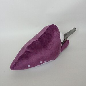 Puede incluir: Juguete de peluche de color p&uacute;rpura con manchas blancas y un tallo gris. El juguete est&aacute; hecho de material suave y est&aacute; dise&ntilde;ado para parecerse a un vegetal estilizado. El juguete probablemente est&aacute; destinado a ni&ntilde;os.