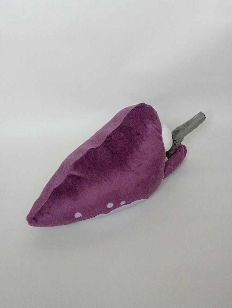 Puede incluir: Juguete de peluche de color p&uacute;rpura con manchas blancas y un tallo gris. El juguete est&aacute; hecho de material suave y est&aacute; dise&ntilde;ado para parecerse a un vegetal estilizado. El juguete probablemente est&aacute; destinado a ni&ntilde;os.