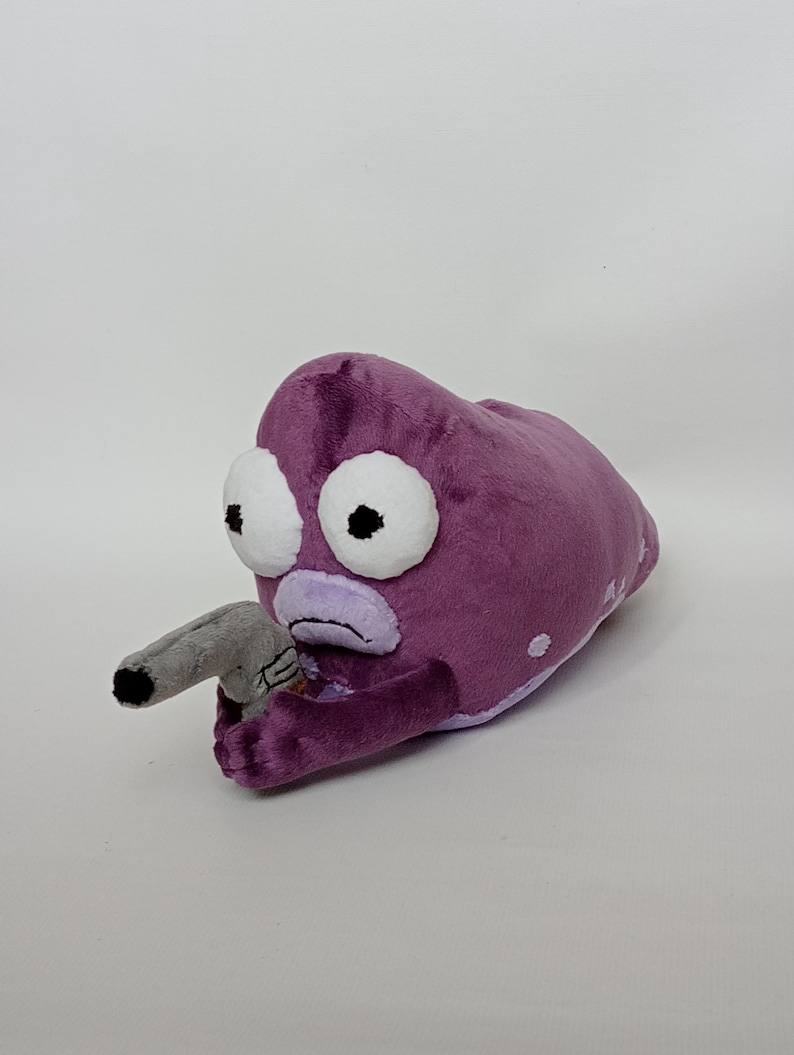 Puede incluir: Juguete de peluche morado con forma de criatura con grandes ojos blancos y un ap&eacute;ndice gris en forma de pistola. El juguete tiene una expresi&oacute;n triste y lunares blancos.