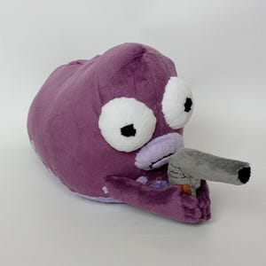 Puede incluir: Un peluche morado con forma de criatura con grandes ojos blancos y una pistola de juguete gris. El juguete tiene una textura suave y difusa, sobre un fondo blanco.