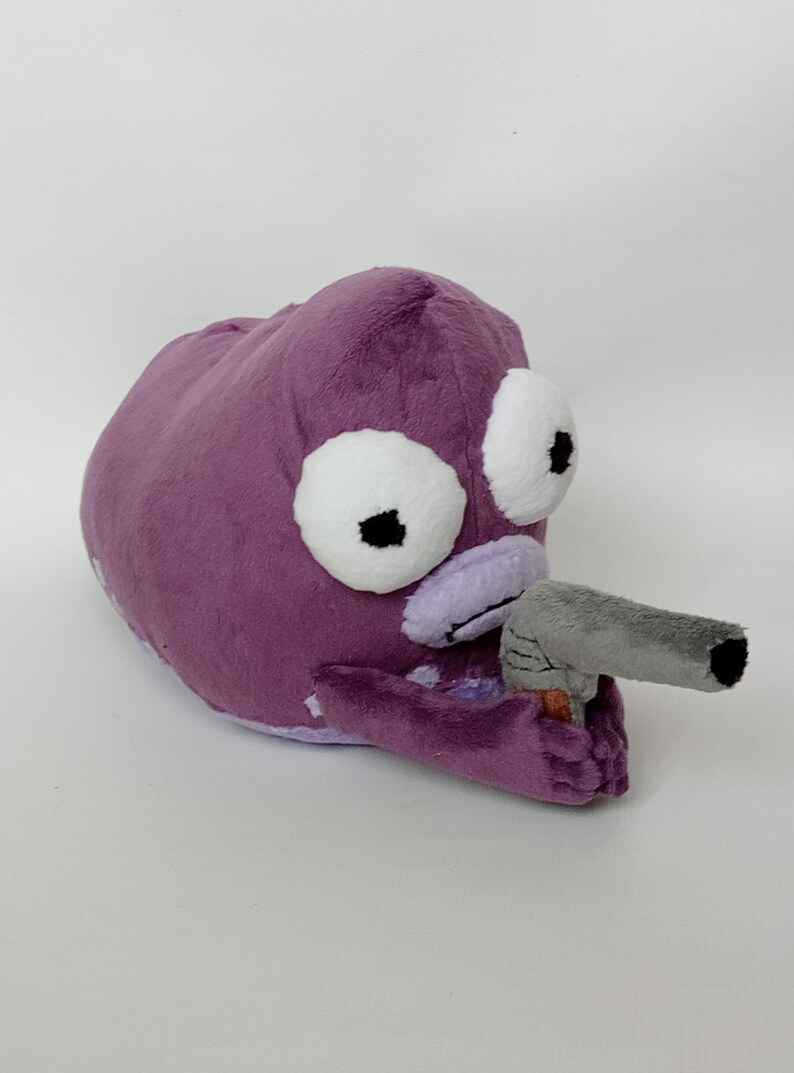 Puede incluir: Un peluche morado con forma de criatura con grandes ojos blancos y una pistola de juguete gris. El juguete tiene una textura suave y difusa, sobre un fondo blanco.