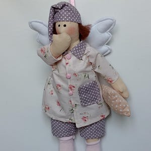 May include: A handmade doll dressed in pajamas with a floral print and polka dot accents. The doll has white wings, a nightcap, and is holding a small pillow. The doll is wearing pink socks and is suspended by a pink ribbon.