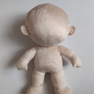 Op de afbeelding: Een beige pluche pop met een rond hoofd, kleine oren en een eenvoudig lichaam. De pop staat op een wit oppervlak.