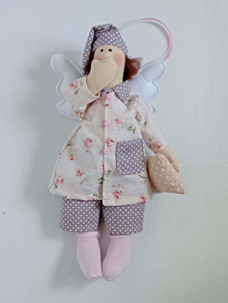 May include: A handmade angel doll wearing pajamas and a nightcap. The doll's pajamas are white with a pink floral pattern, and the pants are gray with white polka dots. The doll has white wings and is holding a small pillow. The doll is approximately 8 inches tall.