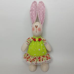 May include: A handmade stuffed bunny doll with long pink polka-dot ears. The bunny wears a green dress with white star accents and a red floral skirt. The doll has a cream-colored face with rosy cheeks and a stitched mouth.