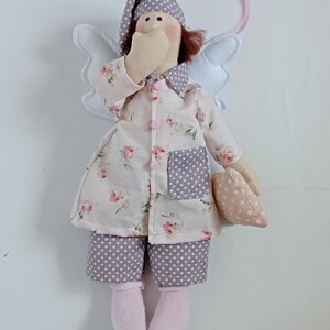 May include: A handmade angel doll wearing pajamas and a nightcap. The doll's pajamas are white with a pink floral pattern, and the pants are gray with white polka dots. The doll has white wings and is holding a small pillow.