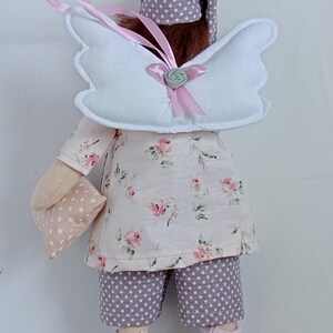 May include: A handmade doll dressed in a floral print top and polka dot shorts. The doll has white angel wings, a matching nightcap, and holds a small pillow. Pink socks and a pink ribbon complete the whimsical design.