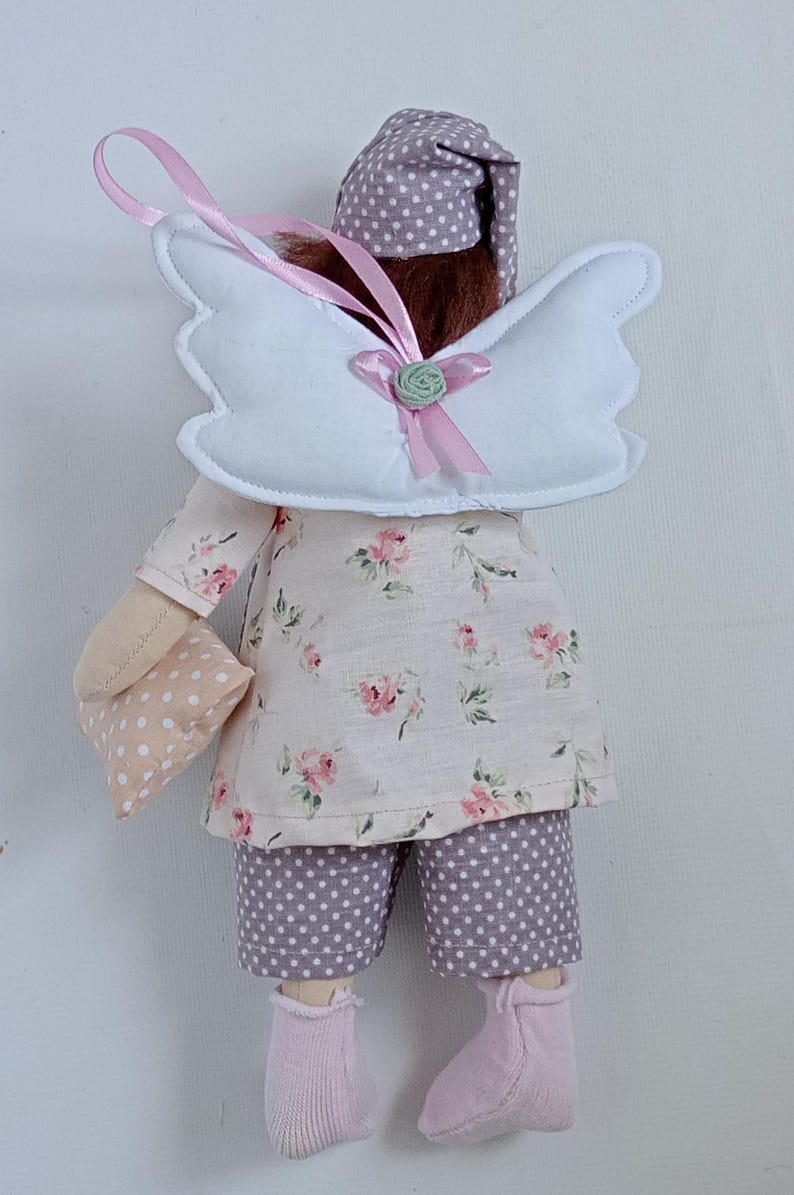May include: A handmade doll dressed in a floral print top and polka dot shorts. The doll has white angel wings, a matching nightcap, and holds a small pillow. Pink socks and a pink ribbon complete the whimsical design.