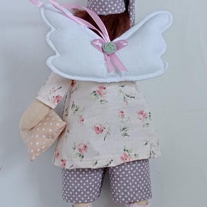 May include: A handmade doll dressed in a floral print top and polka dot shorts. The doll has white angel wings, a matching nightcap, and holds a small pillow. Pink socks and a pink ribbon complete the whimsical design.