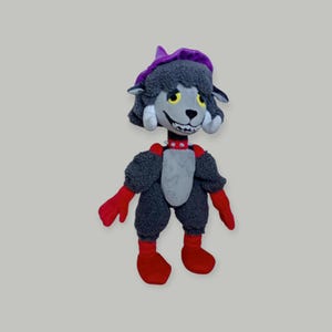 Puede incluir: Un peluche con forma de criatura parecida a un caniche, con un sombrero morado, pelaje gris y detalles rojos. El juguete tiene una cara sonriente con ojos amarillos, un collar rojo, guantes rojos y botas rojas.