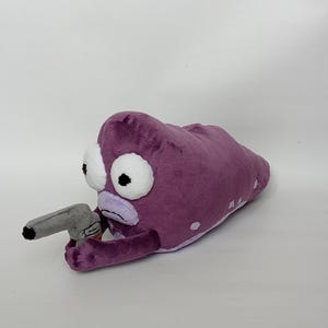 Puede incluir: Juguete de peluche con forma de criatura alien&iacute;gena morada con grandes ojos blancos y una pistola de juguete gris. El peluche tiene manchas blancas y una boca de color lila claro.