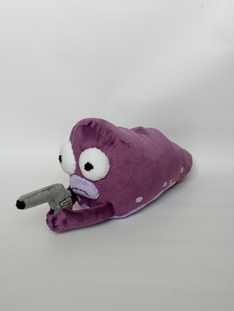 Puede incluir: Juguete de peluche con forma de criatura alien&iacute;gena morada con grandes ojos blancos y una pistola de juguete gris. El peluche tiene manchas blancas y una boca de color lila claro.