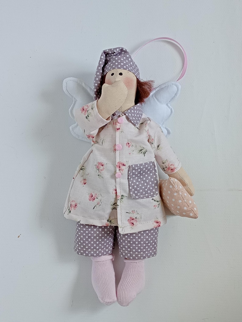 May include: A handmade angel doll wearing a floral print nightgown, polka dot shorts, and a matching nightcap. The doll has white wings, pink socks, and is holding a small pillow. A pink ribbon is attached for hanging.