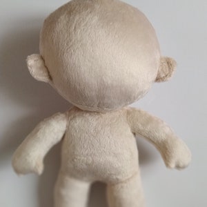 Op de afbeelding: Een effen witte pluche pop met een rond hoofd en kleine oren. De pop staat op een wit oppervlak.