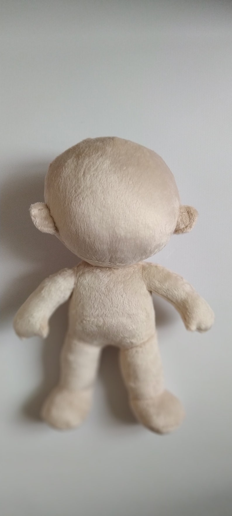 Op de afbeelding: Een effen witte pluche pop met een rond hoofd en kleine oren. De pop staat op een wit oppervlak.
