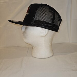 K&ouml;nnte beinhalten: Schwarze Trucker-M&uuml;tze mit Mesh-R&uuml;ckseite und flachem Schirm. Auf der Vorderseite der M&uuml;tze befindet sich ein rot-wei&szlig;es Emblem. Die M&uuml;tze wird auf einem wei&szlig;en Schaumstoffkopf pr&auml;sentiert.