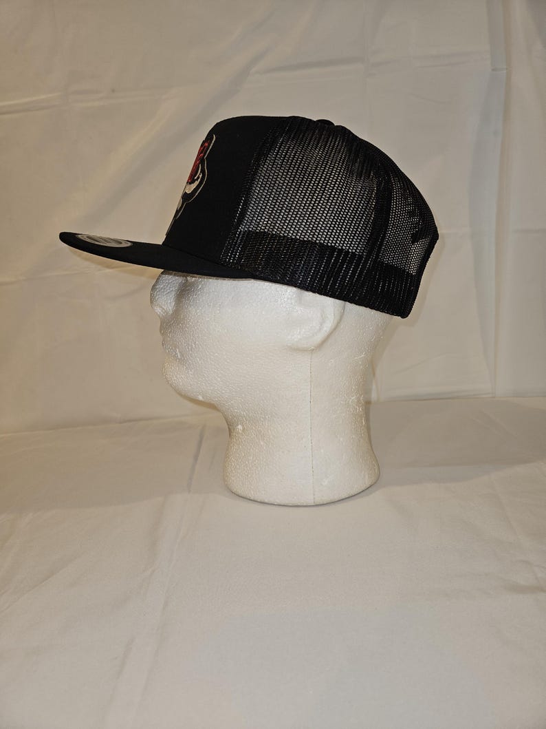 K&ouml;nnte beinhalten: Schwarze Trucker-M&uuml;tze mit Mesh-R&uuml;ckseite und flachem Schirm. Auf der Vorderseite der M&uuml;tze befindet sich ein rot-wei&szlig;es Emblem. Die M&uuml;tze wird auf einem wei&szlig;en Schaumstoffkopf pr&auml;sentiert.