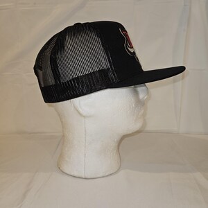 K&ouml;nnte beinhalten: Schwarze Trucker-M&uuml;tze mit Mesh-R&uuml;ckseite und gebogenem Schirm. Auf der Vorderseite der M&uuml;tze befindet sich ein rot-wei&szlig;es Grafikdesign. Die M&uuml;tze wird auf einem wei&szlig;en Schaufensterkopf pr&auml;sentiert.