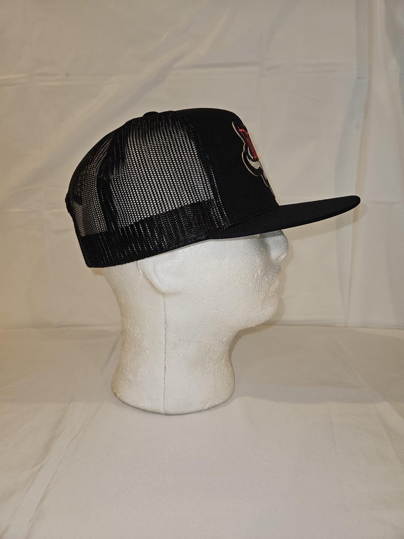 K&ouml;nnte beinhalten: Schwarze Trucker-M&uuml;tze mit Mesh-R&uuml;ckseite und gebogenem Schirm. Auf der Vorderseite der M&uuml;tze befindet sich ein rot-wei&szlig;es Grafikdesign. Die M&uuml;tze wird auf einem wei&szlig;en Schaufensterkopf pr&auml;sentiert.