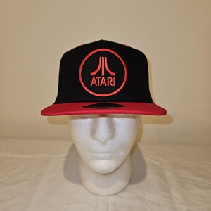 May include: Black and red Atari baseball cap. The cap features a red brim and a black crown with the Atari logo embroidered in red within a red circle. The hat is displayed on a white mannequin head.