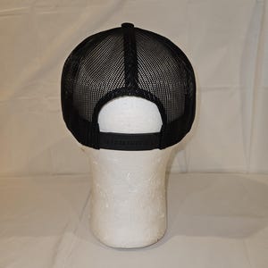K&ouml;nnte beinhalten: Schwarze Trucker-M&uuml;tze mit Mesh-R&uuml;ckseite und verstellbarem Schnappverschluss. Die M&uuml;tze wird auf einem wei&szlig;en Schaufensterkopf pr&auml;sentiert, der das Design und die Passform zeigt. Der Schirm ist schwarz.