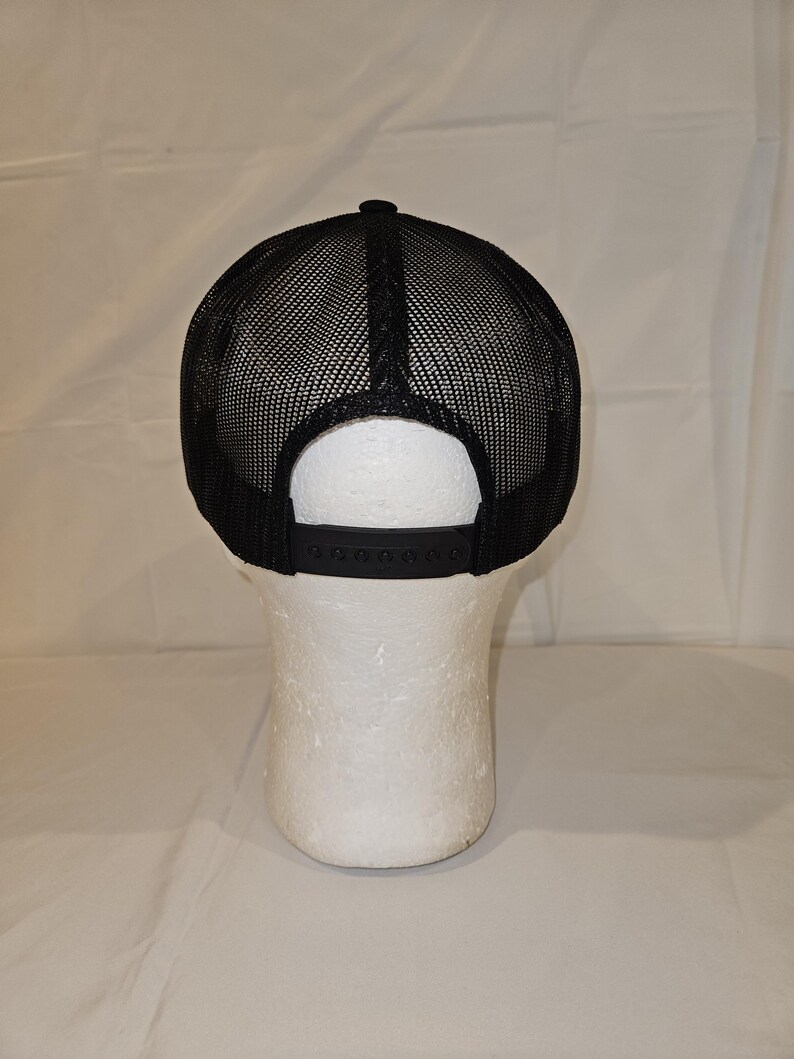 K&ouml;nnte beinhalten: Schwarze Trucker-M&uuml;tze mit Mesh-R&uuml;ckseite und verstellbarem Schnappverschluss. Die M&uuml;tze wird auf einem wei&szlig;en Schaufensterkopf pr&auml;sentiert, der das Design und die Passform zeigt. Der Schirm ist schwarz.